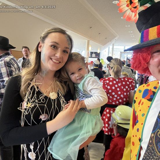 Eine lächelnde Frau in einem Kostüm mit Gesichtsbemalung hält ein Kind in einem grünen Kleid bei einer Kostümveranstaltung, mit einem Clown und anderen in Kostümen im Hintergrund.