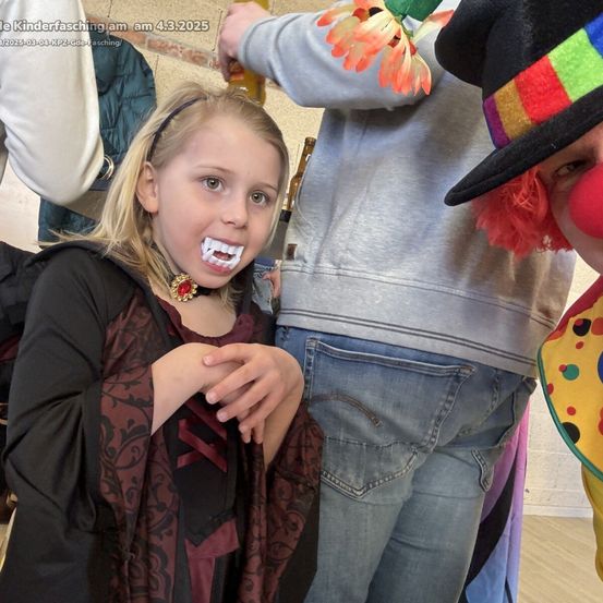 Ein Mädchen in einem Vampirkostüm posiert für ein Foto mit einem Clown im Hintergrund. Ein Mann in Grau hält eine Bierflasche.