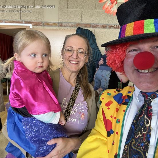 Eine Frau hält ein junges Mädchen in Rosa und Blau und lächelt einen Clown mit roter Nase an. Sie sind in einem Raum mit einer Ziegelwand.