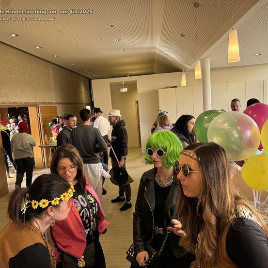 Eine Gruppe von Menschen versammelt sich in einem Raum. Eine Frau mit grünen Haaren und Sonnenbrille unterhält sich mit einer anderen Frau. Rechts sind Luftballons und eine Frau mit einer gelben Blumenkrone schaut sich etwas an.