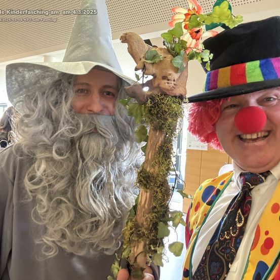 Zwei Personen in fantasievollen Kostümen, einer als Gandalf und der andere als Clown, lächeln für ein Foto. Sie halten einen mit Moos und Blumen geschmückten Stab.