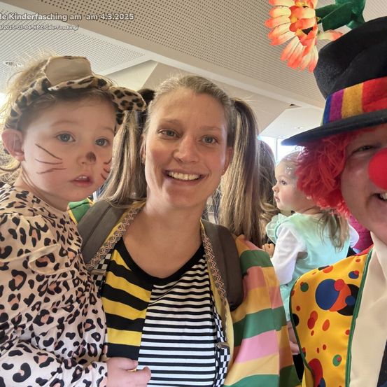 Eine Frau und ein Kind in einem Leoparden-Kostüm lächeln in die Kamera. Hinter ihnen lächeln auch ein weiteres Kind und ein Clown. Der Hintergrund scheint ein Innenraum zu sein.