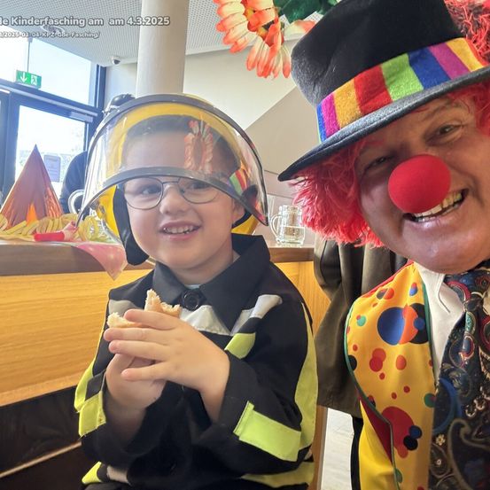 Ein junger Junge in einem Feuerwehrhelm, der ein Sandwich in der Hand hält, lächelt neben einem Clown mit roter Nase, beide posieren für ein Foto. Dahinter steht eine Theke mit bunten Dekorationen.
