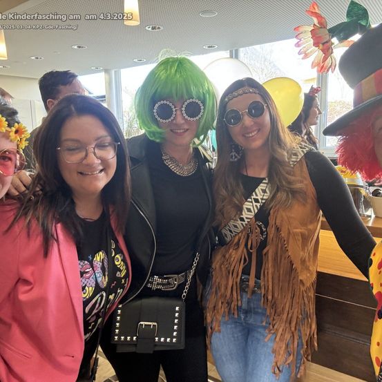 Vier Frauen in bunten, lebendigen Kostümen und Accessoires, die lächelnd für ein Foto in einer Party-ähnlichen Umgebung posieren. Sie stehen eng beieinander, mit Luftballons und Dekorationen im Hintergrund.