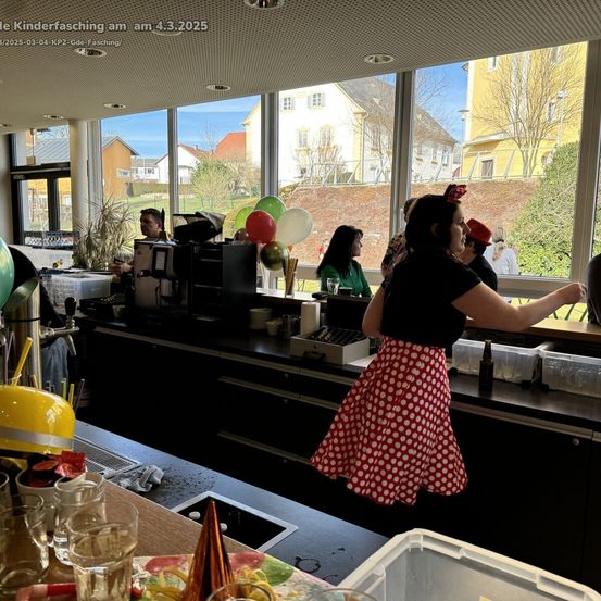Eine Frau in einem Tupfenrock steht hinter einer Bartheke und schaut auf einen Mann mit einer Kaffeemaschine. Auf der Theke stehen Gläser, Tassen und Flaschen. Vor dem Fenster sind Menschen und Ballons zu sehen. Ein Gebäude mit einem Zaun ist in der Ferne sichtbar.