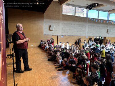 Ein älterer Mann in einem roten Weste und schwarzen Hosen steht mit einem Mikrofon auf der Bühne und spricht eine große Gruppe von Kindern in Kostümen an, die auf dem Boden in einem Schulsaal sitzen.