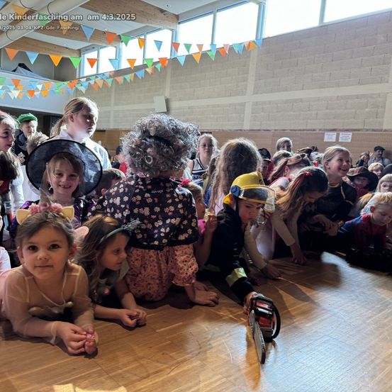 Eine Gruppe von Kindern in Kostümen versammelt sich in einem Saal. Einige sitzen, andere stehen. Eine Person mit einer Kettensägen-Attrappe ist anwesend.