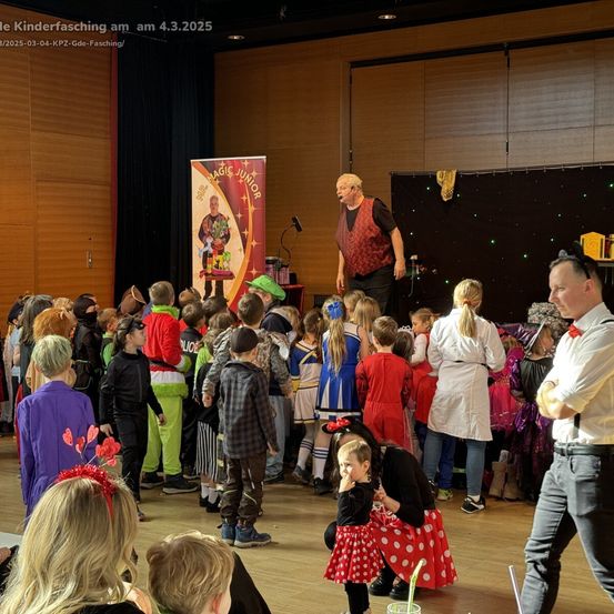 Eine Gruppe von Kindern in Kostümen beobachtet einen Mann auf der Bühne mit einem Banner. Er führt vor, während andere um ihn herumstehen, einige zusehen und andere plaudern.