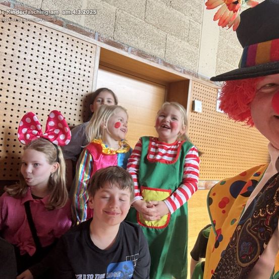 Mehrere Kinder in Kostümen, einschließlich Clown-Make-up, posieren mit einem Clown für ein Foto. Der Hintergrund ist ein Innenraum mit einer Holzwand. Das Datum ist 4.3.2025.