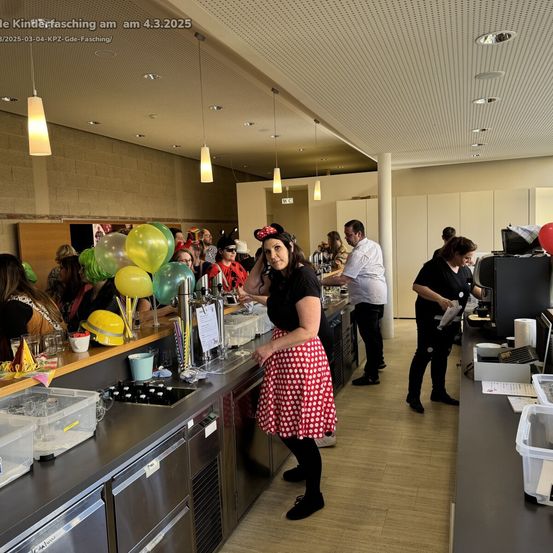 Eine Gruppe von Menschen, darunter eine Frau in einem gepunkteten Kleid, steht an einer Bartheke in einem Restaurant mit Luftballons und Gegenständen auf der Theke.