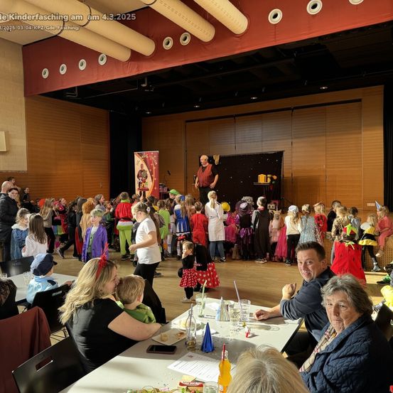 Eine Gruppe von Menschen ist in einem Theater versammelt. Viele Kinder sind in Kostümen gekleidet. Ein Mann steht auf der Bühne. Menschen sitzen an Tischen, einige mit Kindern.