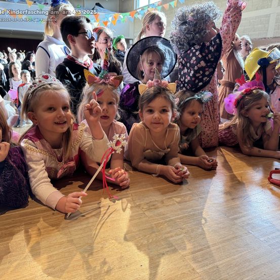 Kinder in Kostümen posieren für ein Foto auf einem Holzboden, einige tragen Stirnbänder und halten Requisiten. Eine Person in einem Piratenkostüm steht hinter ihnen.