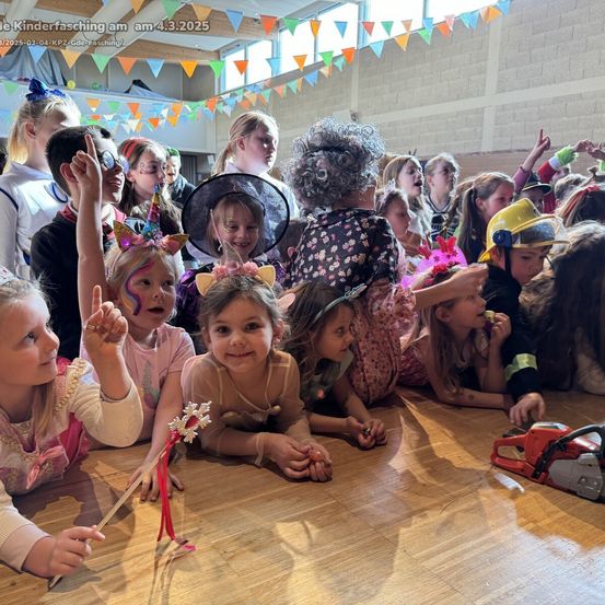 Eine Gruppe von Kindern versammelt sich auf einem Holzboden, einige liegen und einige stehen. Sie sind alle in bunten Kostümen und mit Gesichtsbemalung gekleidet. Ein Kettensäge liegt auf dem Boden.