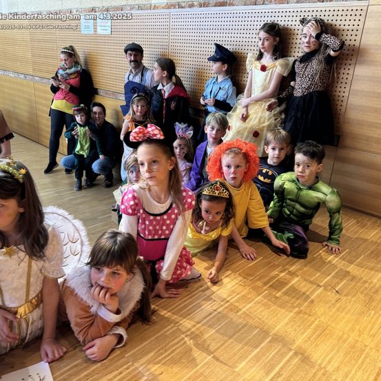 Eine Gruppe von Kindern in verschiedenen Kostümen, darunter Superhelden und Märchenfiguren, posiert für ein Foto auf einem Holzboden.