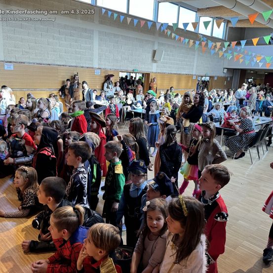 Eine große Menge verkleideter Kinder versammelt sich in einer Halle für einen Kinderfasching am 4. März 2025. Einige sitzen, andere stehen, viele tragen Masken. Im Hintergrund sind Tische und Stühle aufgestellt.