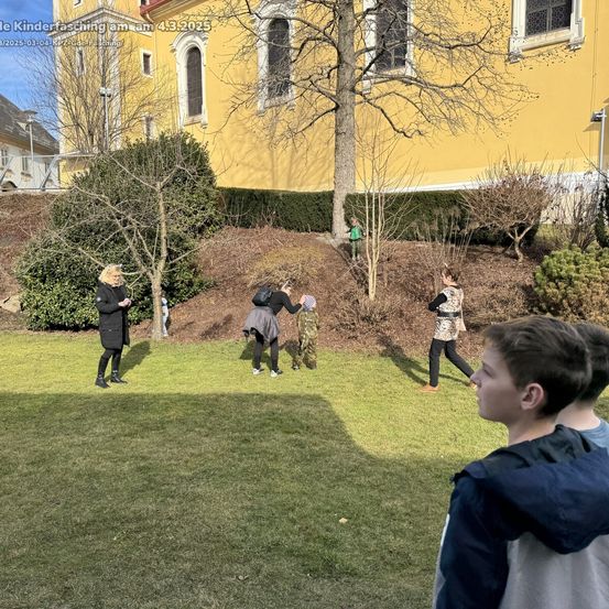 Eine Gruppe von Menschen spielt draußen in der Nähe eines gelben Gebäudes. Sie sind auf einem Rasen mit Bäumen und Büschen. Ein Junge in einem grünen Shirt steht in der Nähe eines Baumes.