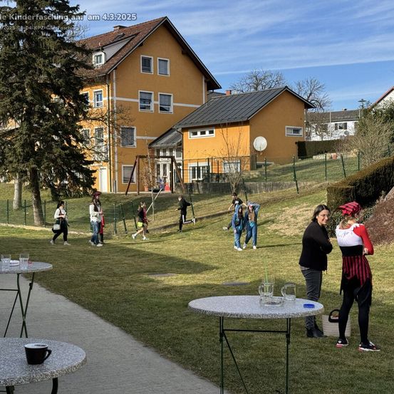 Eine Gruppe von Menschen versammelt sich auf einem Rasenhügel mit Häusern im Hintergrund. Einige gehen, während andere um Tische stehen. Eine Frau in einem roten Kopftuch spricht mit einer Frau in Schwarz.