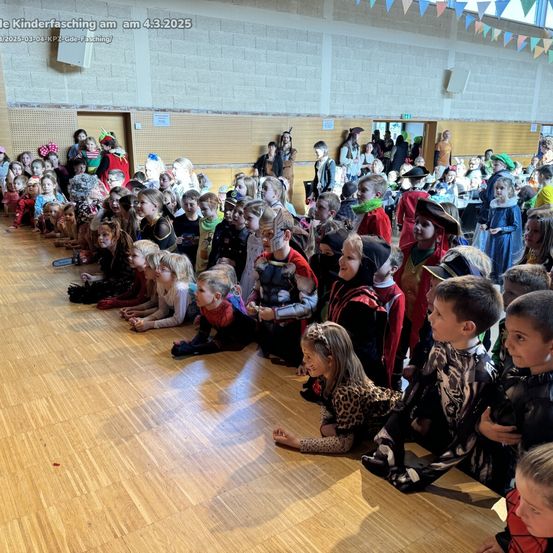 Eine große Gruppe von Kindern in Kostümen sitzt auf einem Holzboden. Viele sind als Superhelden, Piraten und andere Charaktere verkleidet. Die Kinder schauen in verschiedene Richtungen. Das Bild hat einen Wasserzeichen mit Datum und Text.