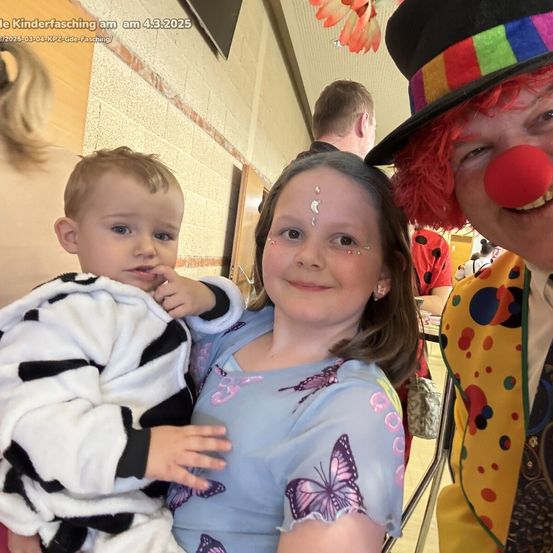 Ein Clown mit roter Nase posiert mit einem Mädchen und einem Jungen in einem Flur, wahrscheinlich bei einer Karnevalsveranstaltung.