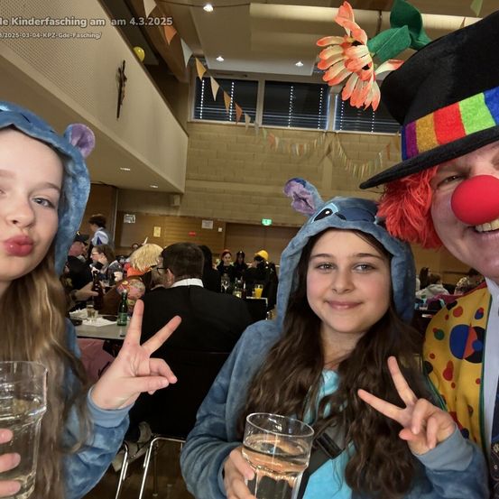 Drei Personen in Kostümen posieren für ein Foto. Zwei Mädchen tragen blaue Kapuzenpullover mit Mausohren, eines hält ein Glas Wasser. Der Junge in der Mitte trägt ein Clownskostüm mit roter Nase. Sie machen Friedenszeichen.