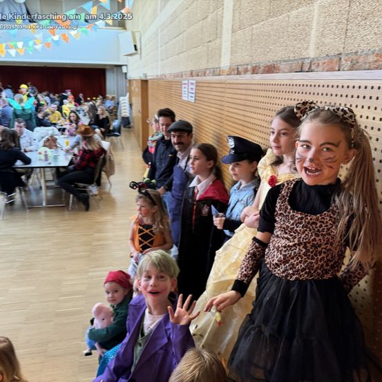 Eine Gruppe von Kindern in Kostümen posiert für ein Foto in einer Halle, mit Erwachsenen und Tischen im Hintergrund. Das Bild hat einen Wasserzeichen in der oberen linken Ecke.