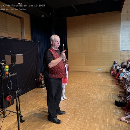 Ein Mann steht auf der Bühne mit einem Mikrofon und spricht vor einer Gruppe von Kindern, die vor ihm auf dem Boden sitzen.