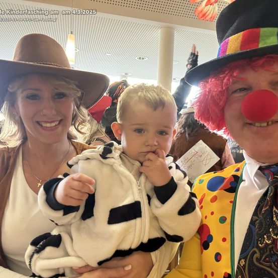 Eine Familie posiert für ein Foto auf einem Jahrmarkt, wobei eine Frau in einem Hut ein Baby in einer Kuh-Kostüm hält und ein Clown neben ihnen steht.