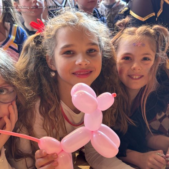 Drei Mädchen, eines davon hält eine rosa Luftballonfigur, lächeln in die Kamera. Das Mädchen in der Mitte trägt ein rotes Band. Im unscharfen Hintergrund sind andere Kinder zu sehen, möglicherweise auf einem Karneval.