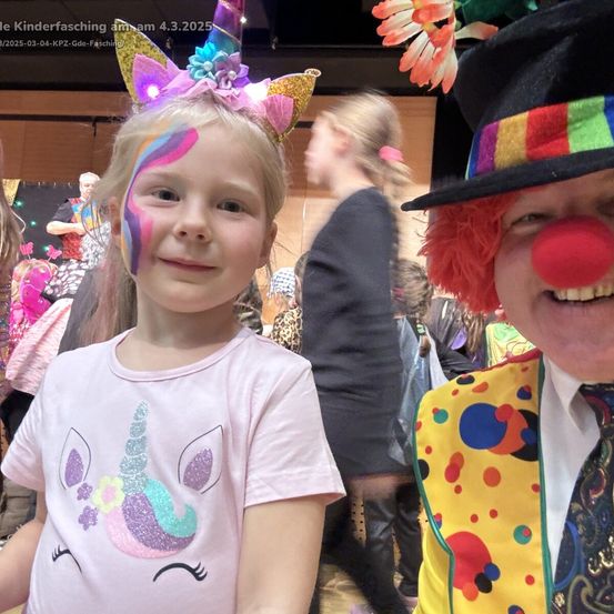 Ein junges Mädchen mit Einhorn-Gesichtsbemalung und ein Clown in einem farbenfrohen Hut. Sie lächeln für die Kamera auf einem Kinderkarneval.