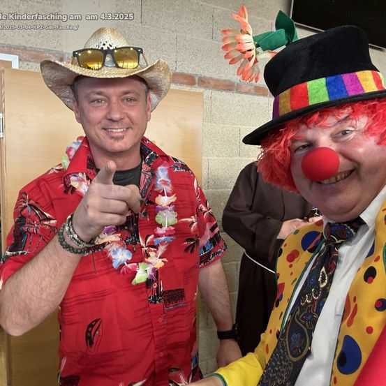 Zwei Männer posieren für ein Foto, einer in einem Cowboyhut und einer Lei, der andere ein Clown mit roter Nase. Dahinter ein Priester in einem braunen Gewand.