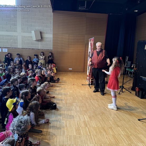 Ein Zauberer führt auf der Bühne vor einer Gruppe von Kindern in Kostümen vor. Die Kinder sitzen auf dem Boden und schauen aufmerksam zu.