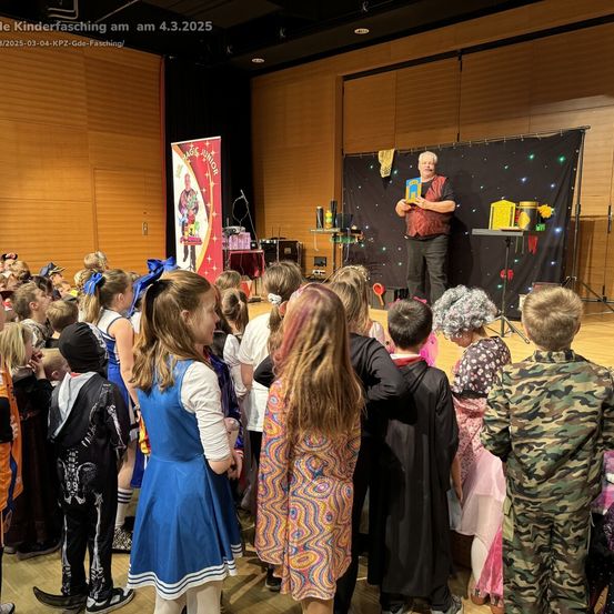 Kinder in Kostümen beobachten einen Magier auf der Bühne. Der Magier hält ein Buch und Gegenstände, hinter ihm befindet sich ein Banner. Die Bühne ist mit einem schwarzen Hintergrund dekoriert.