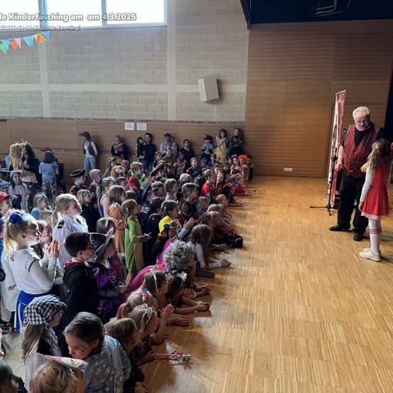 Ein Saal voller Kinder in Kostümen, die einen Mann in einem roten Vest und einen anderen in einem roten Kleid auf der Bühne beobachten.