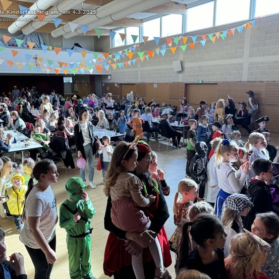 Eine große Menschenmenge aus Kindern und Erwachsenen in verschiedenen Kostümen versammelt sich in einer Halle. Viele halten Schilder und Luftballons hoch, und im Hintergrund sind Tische mit Essen aufgestellt. Die Halle ist mit bunten Bannern geschmückt.