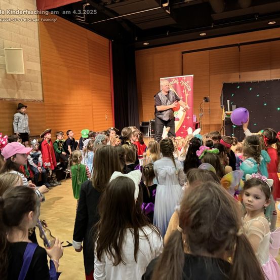 Ein Mann auf der Bühne führt Zaubertricks für ein großes Publikum von Kindern in Kostümen vor. Er hält einen roten Ball und eine lila Ballon. Die Kinder sind um ihn herum versammelt und beobachten ihn aufmerksam. Die Umgebung ist drinnen, mit einem Holzboden und Wänden.