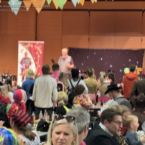 Eine Menschenmenge versammelt sich in einem Veranstaltungsort für einen Kinderkarneval, mit einer Bühne im Vordergrund, auf der ein Magier in einem farbenfrohen Kostüm steht. Bunte Banner und Luftballons sind sichtbar.
