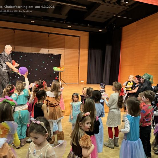 Eine Gruppe von Kindern in verschiedenen Kostümen ist auf einer Bühne versammelt und beobachtet einen Mann, der mit Luftballons performt. Die Umgebung ist in Innenräumen mit Holzwänden und einem schwarzen Hintergrund.