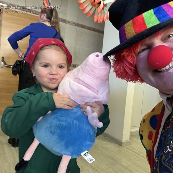 Ein junges Mädchen in einem grünen Pullover hält ein rosa Plüschtier, während es neben einem Clown steht, der einen farbenfrohen Hut trägt. Im Hintergrund steht eine weitere Person in einem blauen Hemd in der Nähe einer Tür.