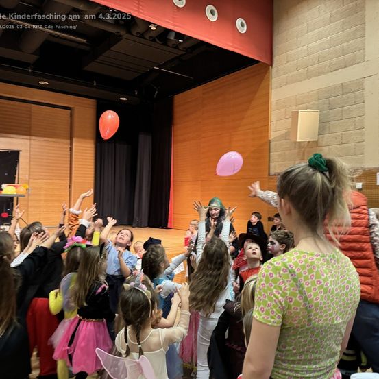 Kinder in Kostümen heben während einer Karnevalsveranstaltung am 4. März 2025 ihre Hände. Eine Person in der Mitte mit grüner Maske wirft Ballons.