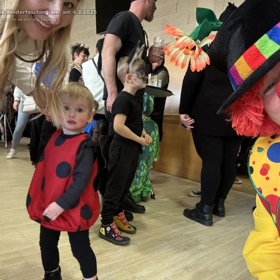 Kinder in Kostümen versammeln sich drinnen. Ein Kleinkind in einem Marienkäferkostüm sticht hervor. Andere sind als Clown und Blume verkleidet. Das Datum ist der 4. März 2025.