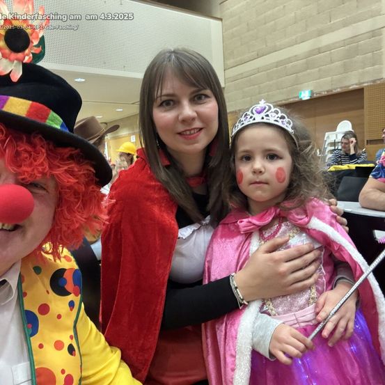 Ein Foto vom Kinderfasching am 4.3.2025 mit einem Clown, einer Frau in einem roten Umhang und einem Mädchen in einer Prinzessinnen-Verkleidung. Das Mädchen hält eine Zauberstange und hat rote Wangen.