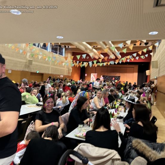 Eine große Menschenmenge, darunter Kinder, versammelte sich am 4. März 2025 zu einem Karnevalsevent in einem Raum. Viele sitzen an Tischen mit Essen, Getränken und Dekorationen. Einige tragen Kostüme. Die Decke hat Wimpelketten.