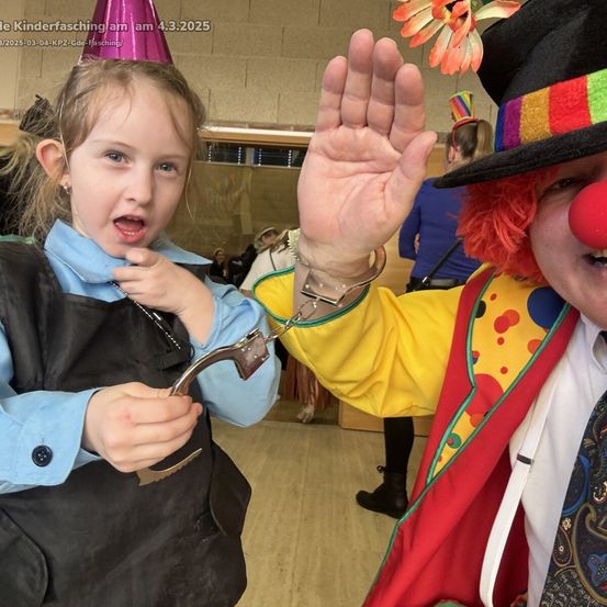 Ein Mädchen in einem Kostüm hält eine Clown-Handschellen, während ein Clown in einem bunten Kostüm eine Hand erhebt. Der Ort scheint eine Kinderfeier zu sein.