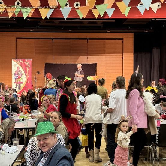 Eine lebhafte Veranstaltung mit einer Menschenmenge, Erwachsenen und Kindern, in Kostümen gekleidet. Ein Mann steht auf der Bühne, und ein Banner ist vorhanden. Tische sind mit Essen und Getränken gedeckt. Die Umgebung erscheint festlich mit Dekorationen und Fahnen.
