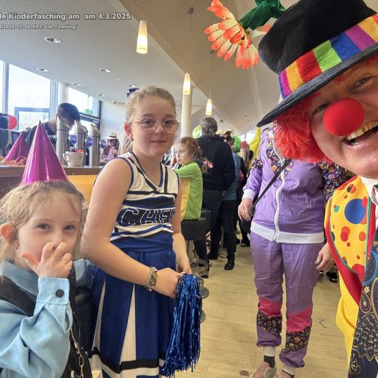 Ein lächelnder Clown macht ein Selfie mit zwei Mädchen, eines in einer Cheerleader-Uniform und das andere in einer blauen Jacke, in einem geschäftigen Bereich mit Partyhüten und Luftballons.
