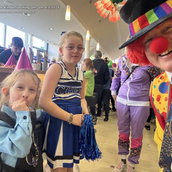 Ein junges Mädchen mit Brille und Pompons steht zwischen zwei anderen Kindern, von denen eines als Clown verkleidet ist. Sie stehen alle in einem überfüllten Raum.