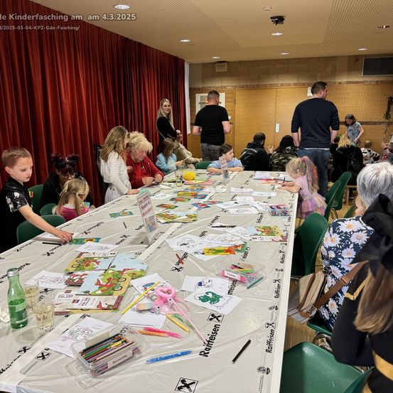 Eine Gruppe von Kindern und Erwachsenen sitzt um einen langen Tisch herum und füllt verschiedene Bilder aus. Sie sind von bunten Stiften und Markern umgeben. Der Raum hat eine rote Vorhangkulisse.