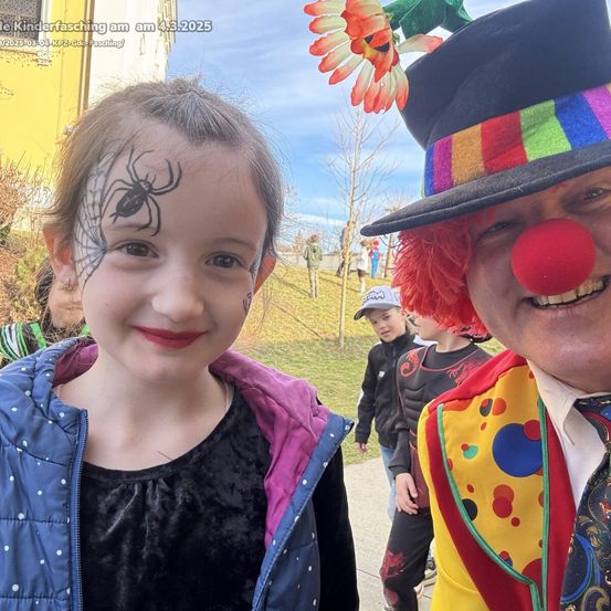 Ein junges Mädchen mit einer Spinnennetz-Gesichtsbemalung und ein Clown mit roter Nase. Sie lächeln in die Kamera, mit Leuten, die im Hintergrund vorbeilaufen.