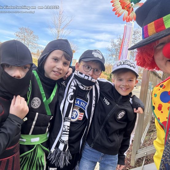 Fünf Jungen in verschiedenen Kostümen posieren für ein Foto. Der Junge in der Mitte trägt eine schwarze Jacke mit weißen Streifen. Der Junge rechts trägt einen Hut. Hinter ihnen steht ein Zaun mit einer Fahne.