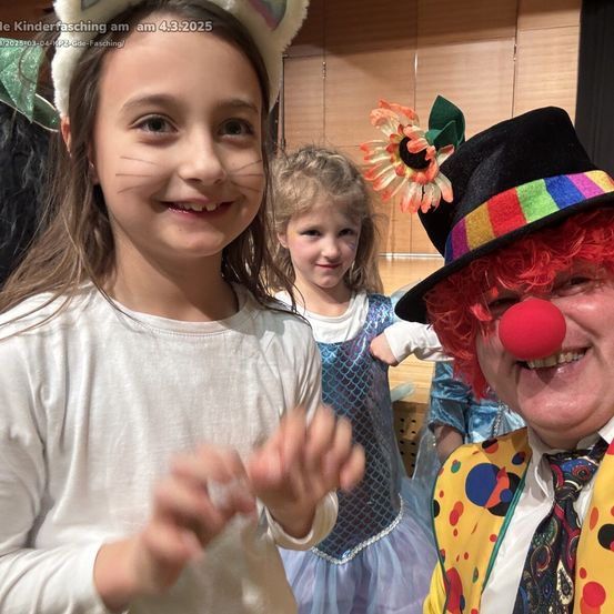 Ein Mädchen in einem Katzenkostüm lächelt neben einem Clown. Dahinter steht ein weiteres Mädchen in einem blauen Kleid. Das Bild ist vom Kinderfasching am 4. März 2025.
